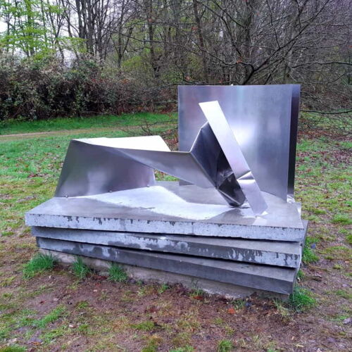 Eine moderne Skulptur aus glänzendem Metall mit kantigen, ineinander verschlungenen Formen steht auf einem gestuften Betonsockel in einem grünen Park. Im Hintergrund sind Bäume und ein Weg zu sehen.